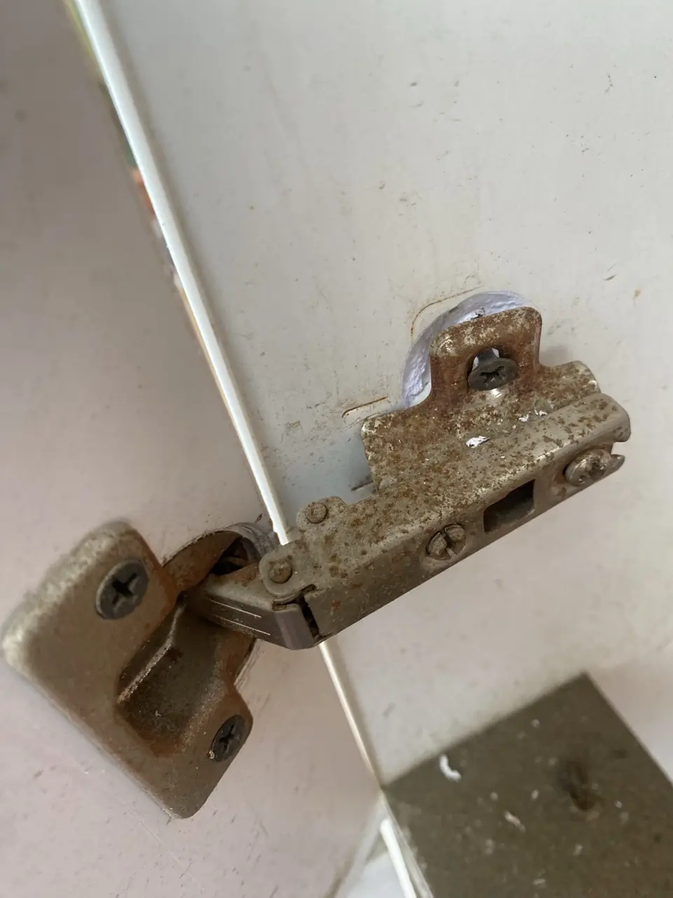 Rusted concealed kitchen cabinet hinge on a white cupboard door before replacement in a Handyman MultiFix repair.